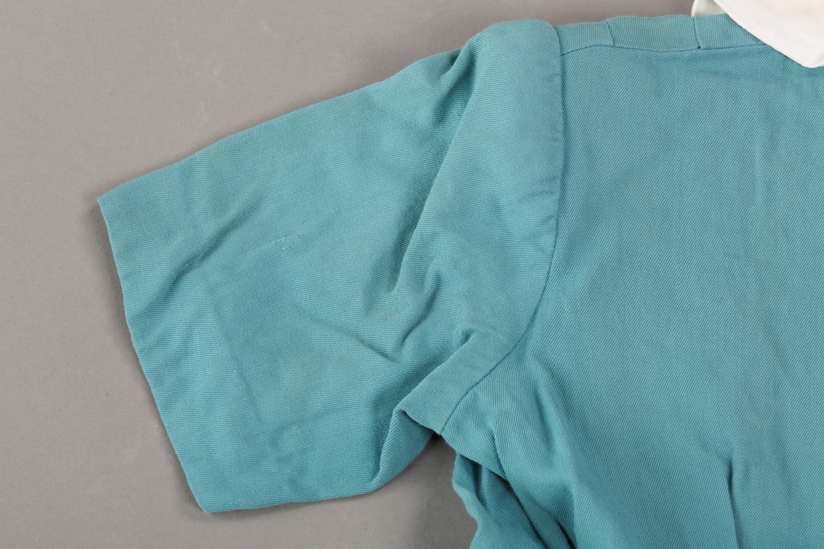 Form: Innsvingt liv, korte ermer, plissert skjørt, fastsydd belte.
Krave.
Bærestykket har vertikale stolper på hver side fra skulder ned til liv, med sydde legg.
Åpning foran med trykknapper. 3 pynteknapper utenpå.