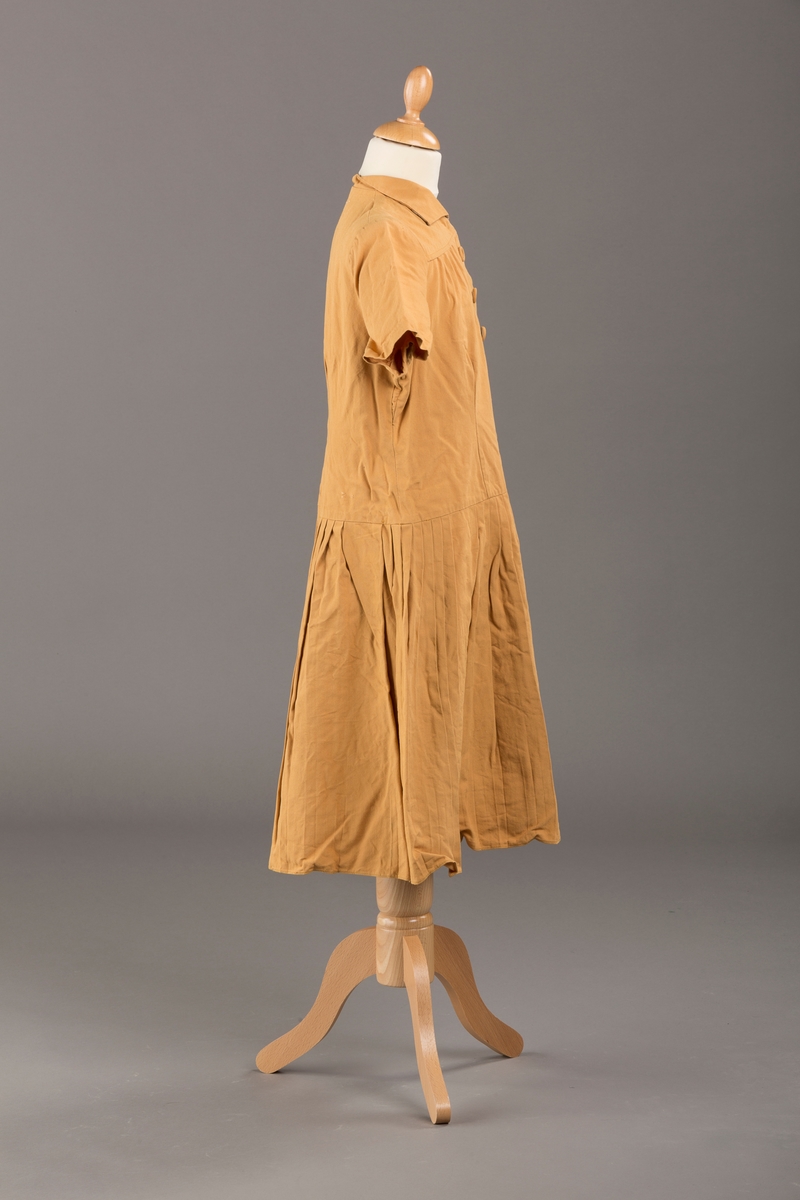 Form: Korte ermer, livet rynket til bærestykke foran, plissert skjørt, tre pynteknapper, en trykknapp, forlomme.