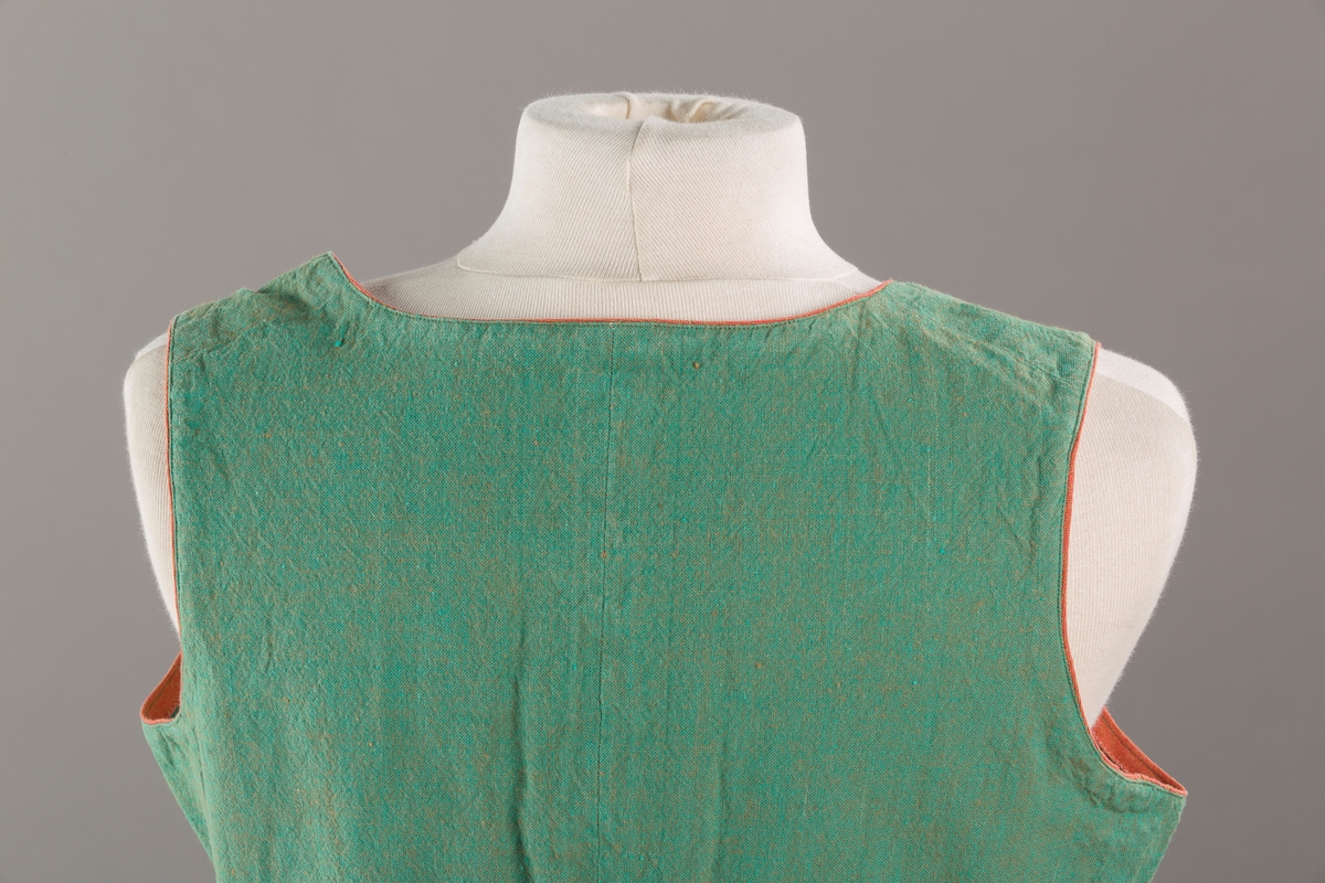 Grønt liv med rustrød kant og fór, seks skjøter. Håndsydd forsterkning i kontrastfarge øverst i skjøtesplittene. Lukkes med tinnhekte.