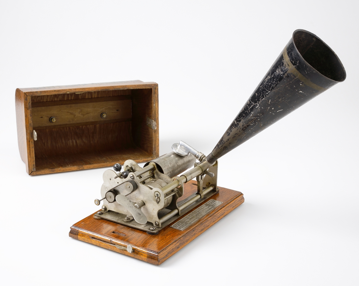Fonograf.
Komplett och fungerande fonograf bestående av träfodral (bottenplatta och lock i trä), tratt i svartlackerad plåt och fonografspelare.