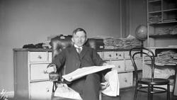 Mann sitter i stol omgitt av gamle papirer eller aviser.