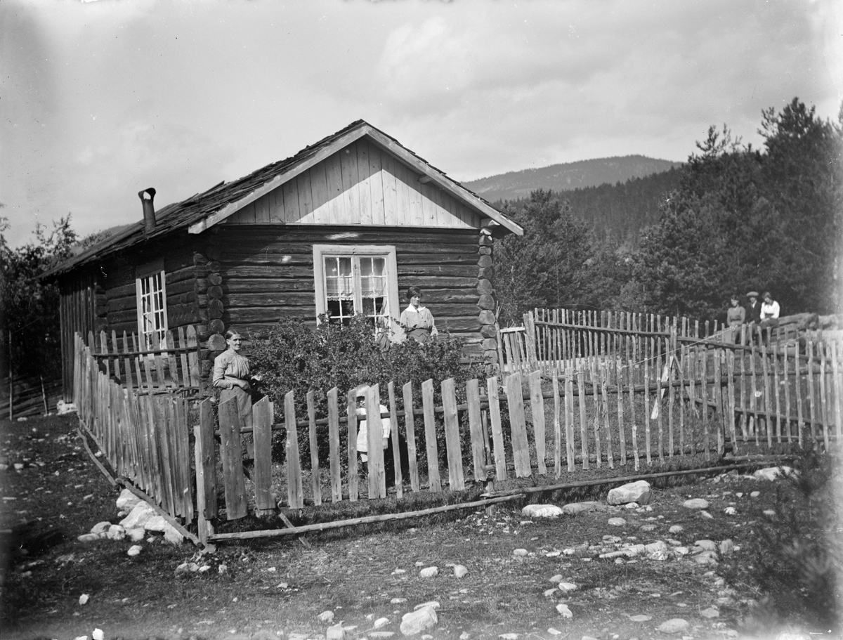 Sperre/Olsen-huset i Tollmoen, Ringebu. Tømret hus med inngjerdet hageflekk. Folk i hagen og på stein i bakgrunn.