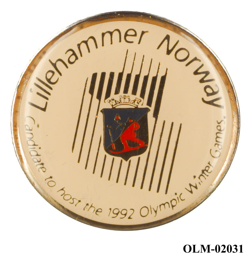 Rundt merke med en tidlig versjon av nordlyslogoen og byvåpenet for Lillehammer kommune. Tekst plassert rundt kanten. Merket har gulnet.