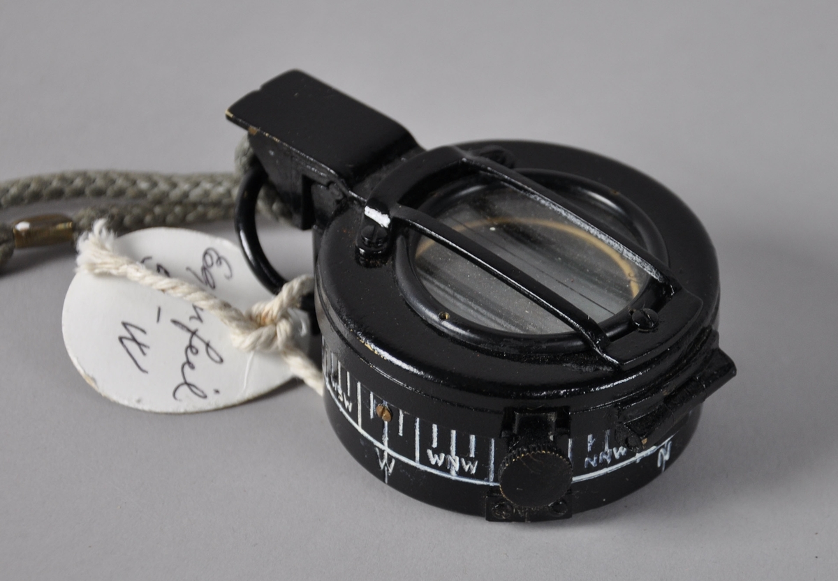 Sort kompass av metall. Til kompasset følger det med en taske av stoff. Tasken har hemper for feste til belte.