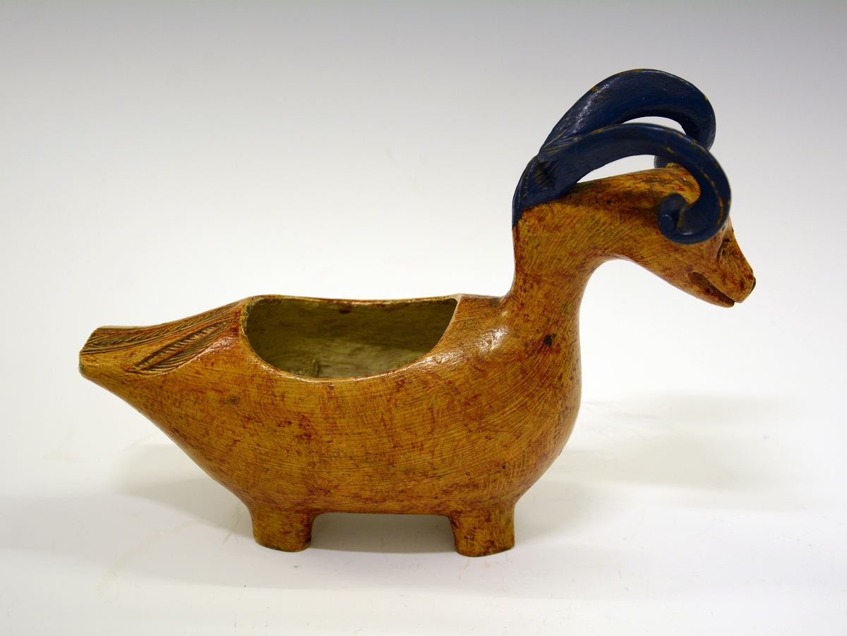 Kane. Fra protokollen: Kane, saak. "ølbøkaar", av træ. Kroppen nærmest mindende om gris + and, med 4 avstumpede ben og avstumpet hale. Hovedet bærer 2 foroverbøiede, fritliggende væderhorn (værhorn), som med sin forreste krumning hviler paa kjæven; hovedet forøvrigt væderlignende. Sen maling, kirketræsfarvet krop, blaa horn, hvit inside.