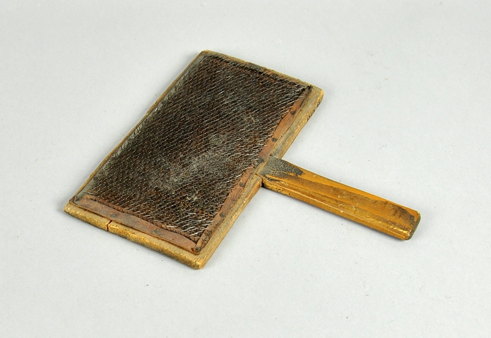 Treplate med håndtak der den ene siden av platen er dekket av lærduk med tettsittende nåler. Treplaten er sprukket.