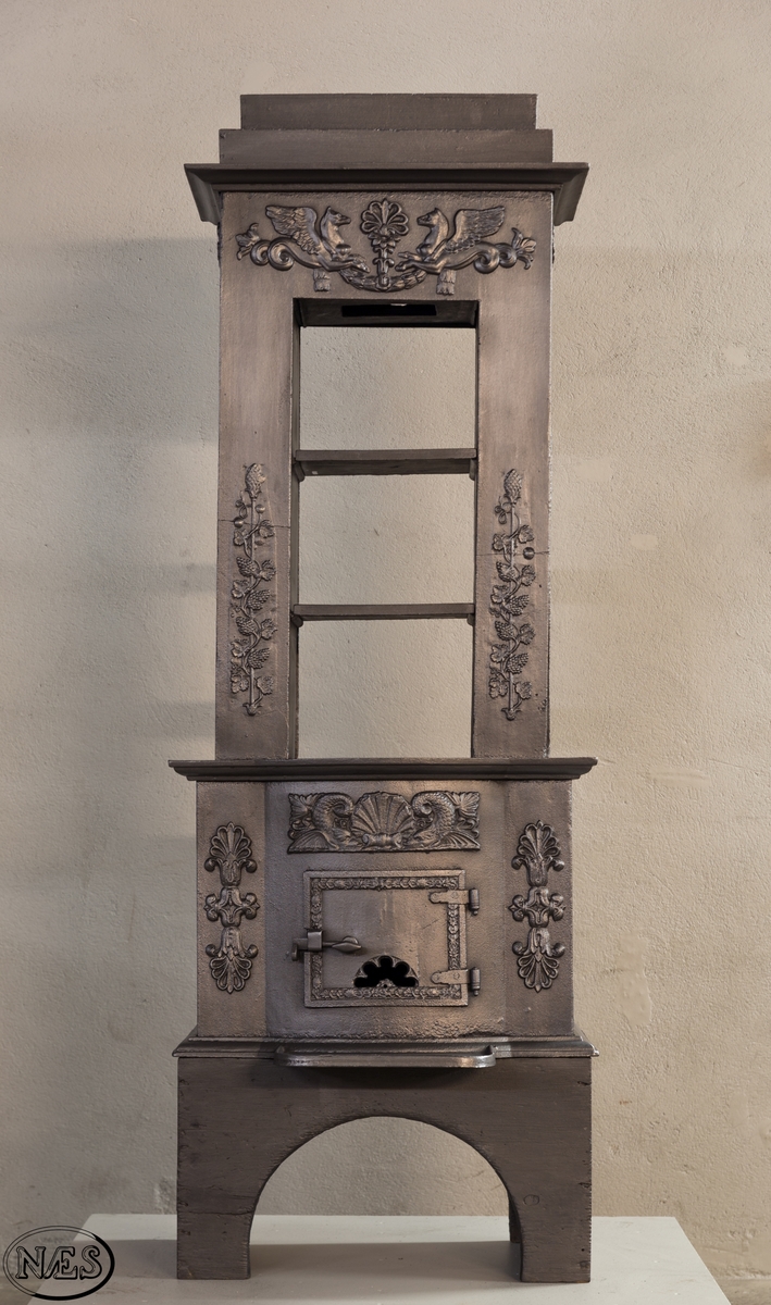 Ovnen består av to avdelinger. Underdelen har et frembuet midtparti med kamskjell flankert av sjødyr på hver side. På kortsidene skjold, løvehode og symmetriske ørner. På bakplate en krukke med symmetriske engler.
Overdelen er høy, firkantet med tredelt åpning. Empire ornamentikk og avtrappet topp. Øverst på frontplaten symetriske fabeldyr (pegasus) vendt mot hverandre.