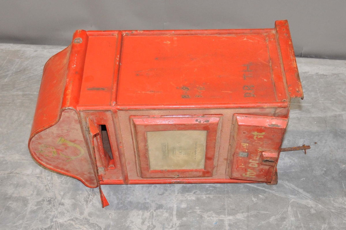 Rød mekanisk tømmepostkasse for tømmeapparat. Med posthornemblem og inntilling for tømmetider.  Malingsslitt. Opplysningsplakat om tømmetider.