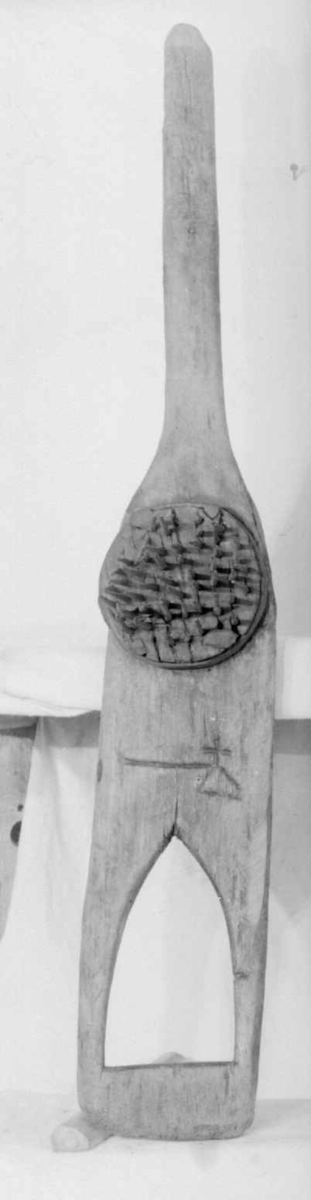 Linhekle som har et håndtak i den ene enden, den andre enden er smal og spiss. På midten av heklen er det en rundt kloss med stålspiker. Det er skåret ut et merke på brettet bestående av en trekant, en strek og et kors.