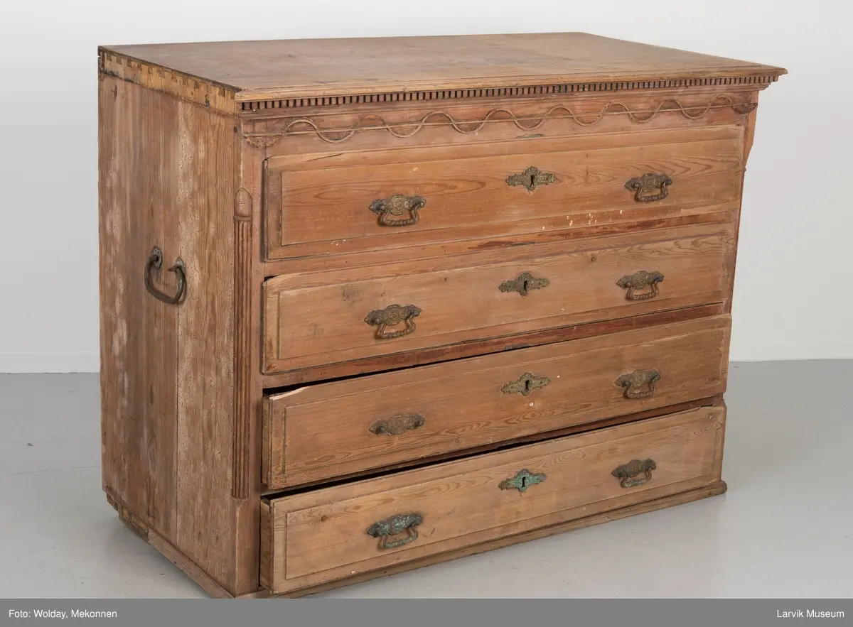 Kommoden har 4 skuffer. 2 beslag for å dra ut, 1 beslag for nøkkelhull, messing. Utskåret dekor under øvre pyntelist "tanngard" rundt på 3 kanter. Øvre skuff utskåret med bølgebånd med rett linje som bryter bølgene. Utskjæring kun i front. To av kommodens hjørner har utskåret søyle. Håndtak på kortsidene for å bære i.