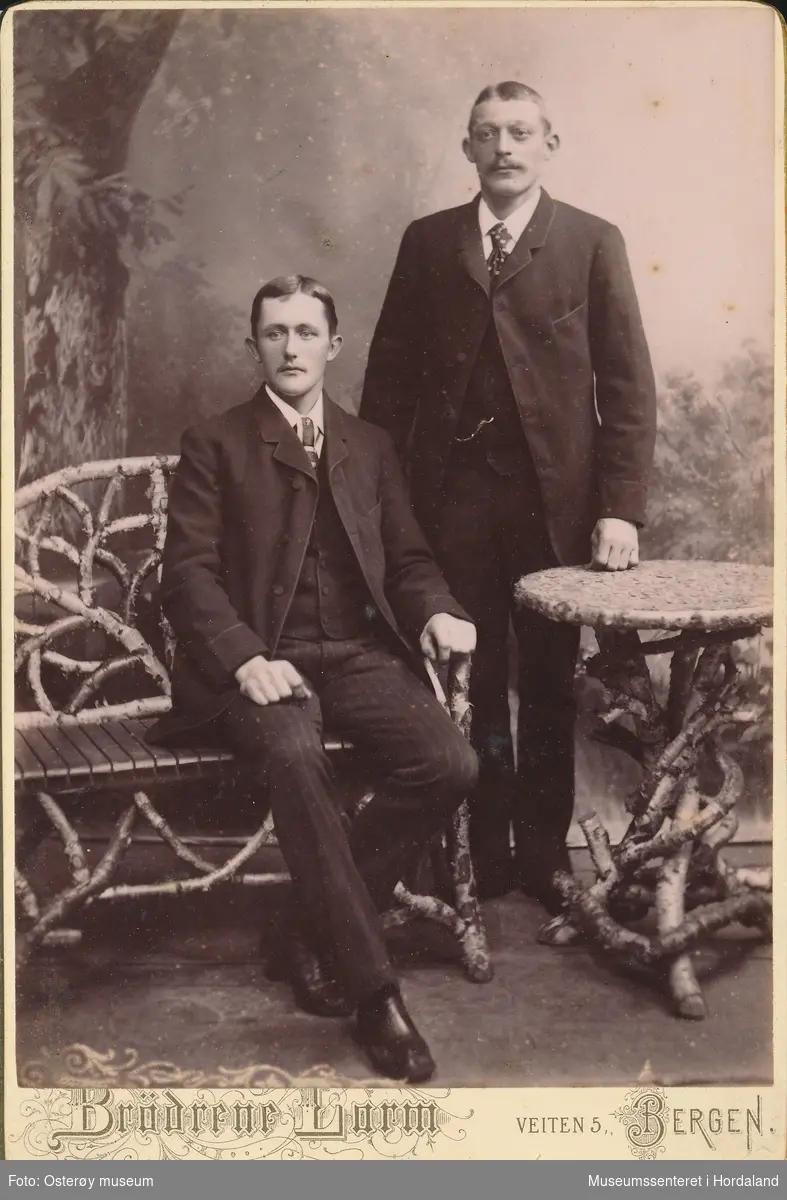 atelierfotografi av to unge menn i  mørke dresser med vest, kvit skjorte og mønstra slips