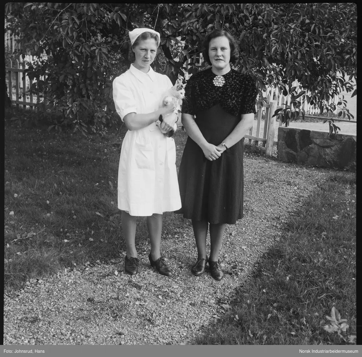 Gruppeportrett, to kvinner stående i hage kledd i uniform og kjole. En av kvinnene har en katt på armen. Elever ved Husmorskolen.