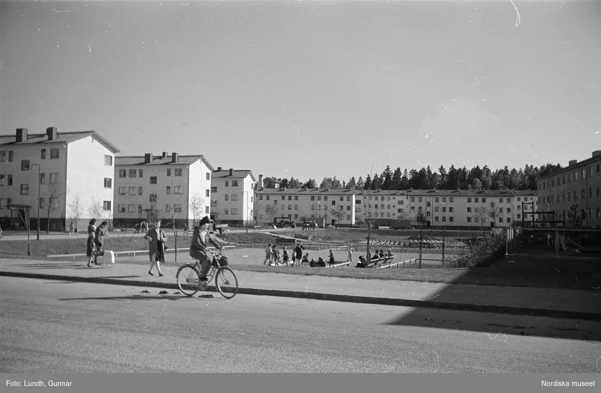 Spångavägen, Bromma, Stockholm. I bakgrunden bostadshus, i förgrunden lekande barn på gård, fotgängare och en kvinna på cykel.