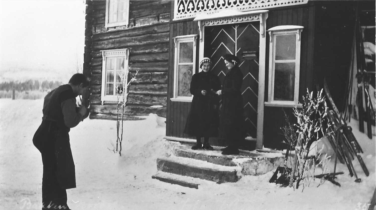 Personer foran bolighus, den ene fotograferer to kvinner på trappa