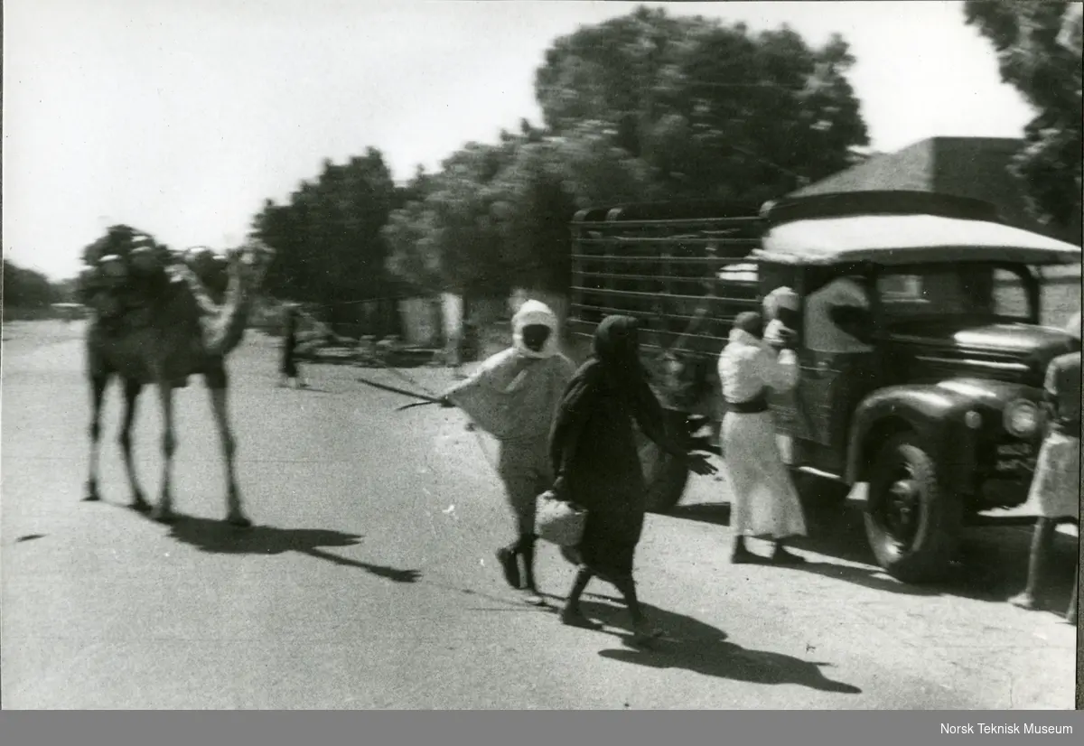 Sudanske kvinner, én leier kamel med last. Til høyre står en Ford 1946-47 lastebil.