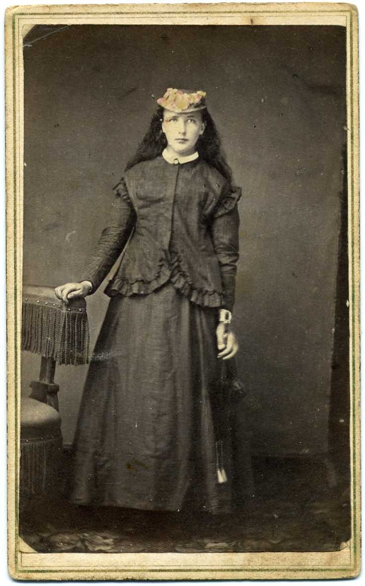 Kabinettsfotografi: okänd ung kvinna med långt hår stående vid ett bord; liten hatt på huvudet och parasoll i handen.