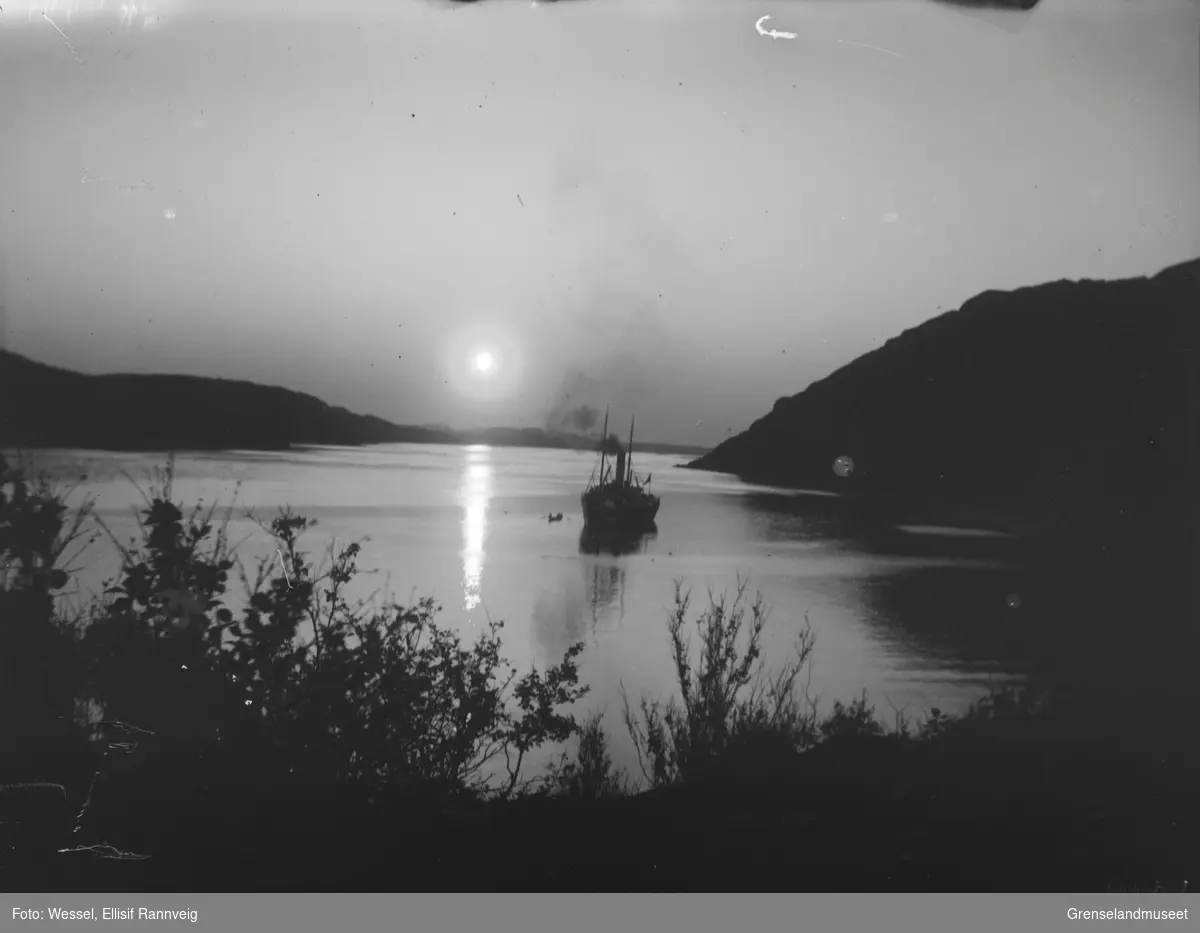 En dampbåt ligger ute på sjøen ved Elvenes, en jolle er på vei ut. Midnattsola skinner.