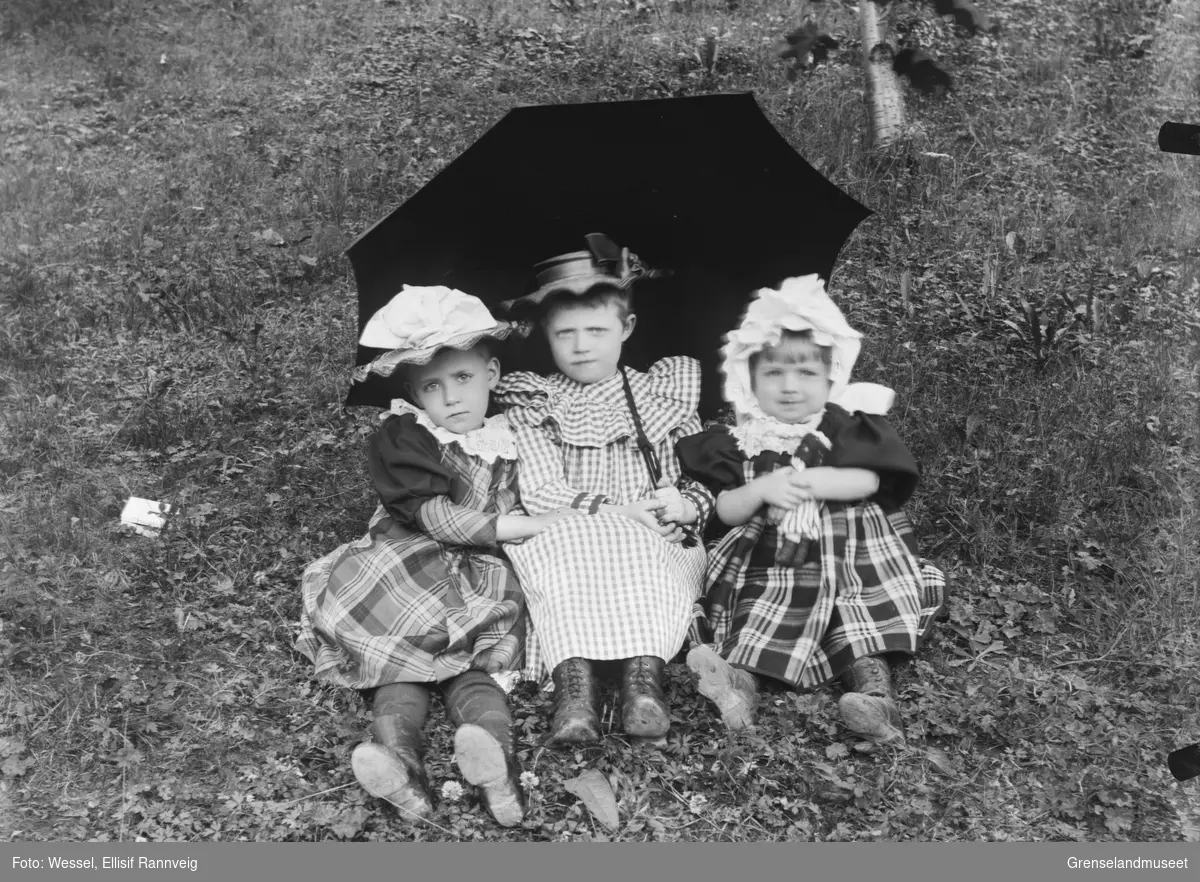 Tre små jenter fotografert sittende i gresset under en paraply.