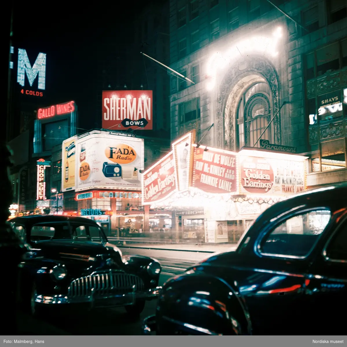 New York, USA. Gata i kvällsbelysning. En biograf visar filmen Golden Earrings. Diverse neonskyltreklam på husfasaderna, parkerade bilar i förgrunden.