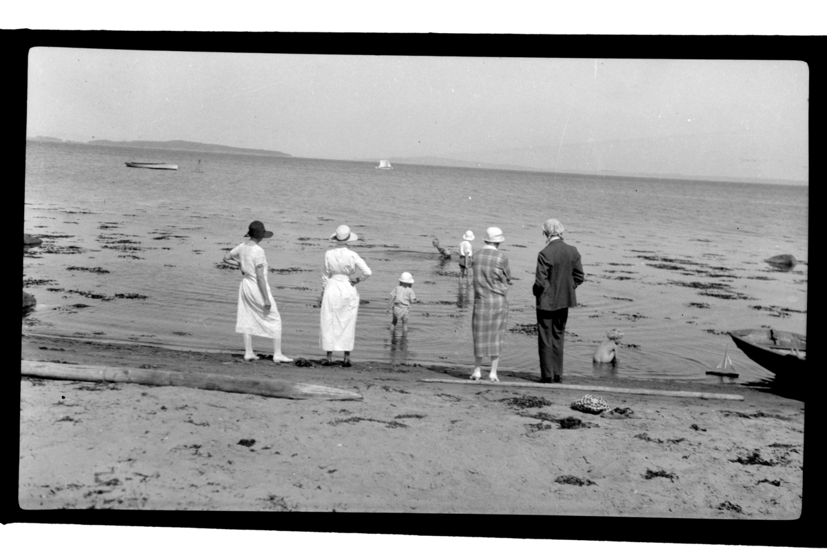 En gruppe voksne og barn på sandstrand. Barna bader. Fotografert ca. 1929.