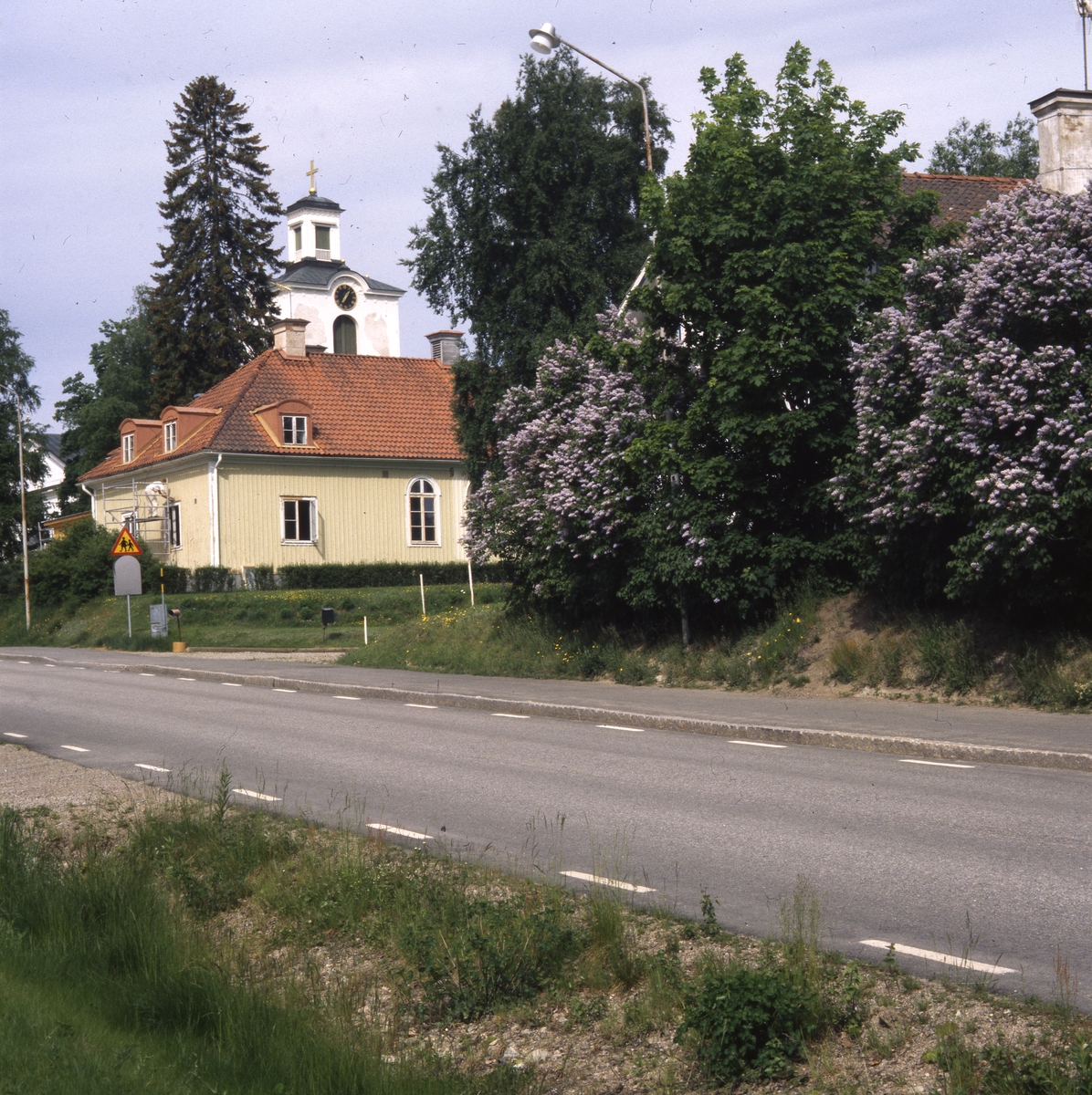Rengsjö församlingshus med kyrkans torn i bakgrunden, sett från vägen, juni 1999.