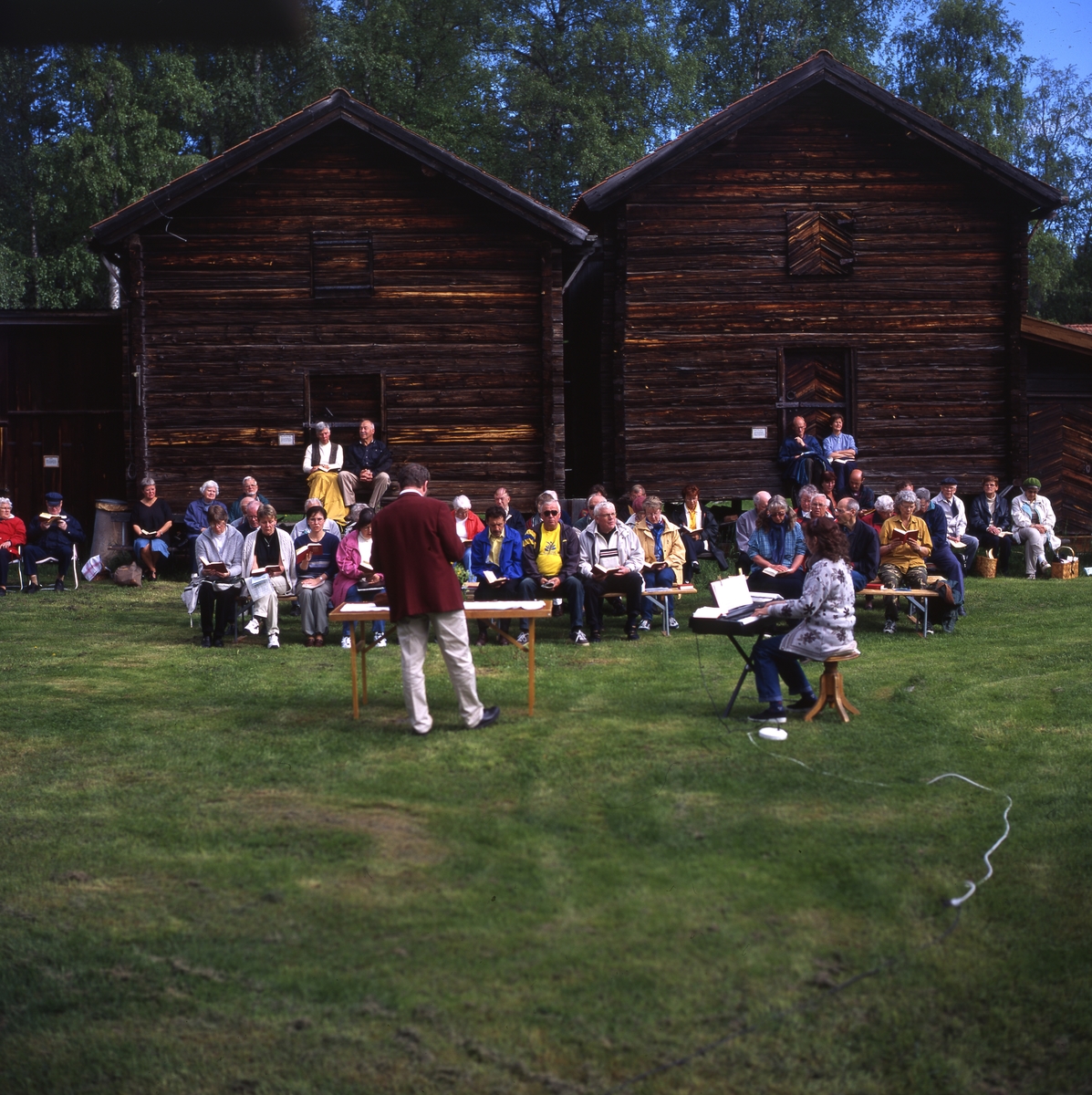 Gökotta i Västerby hembygdsby, Rengsjö 1 juni 2000. Deltagarna sitter på bänkar vid härbrena för att lyssna till sång och musik. En del har med sig matsäckskorgar.