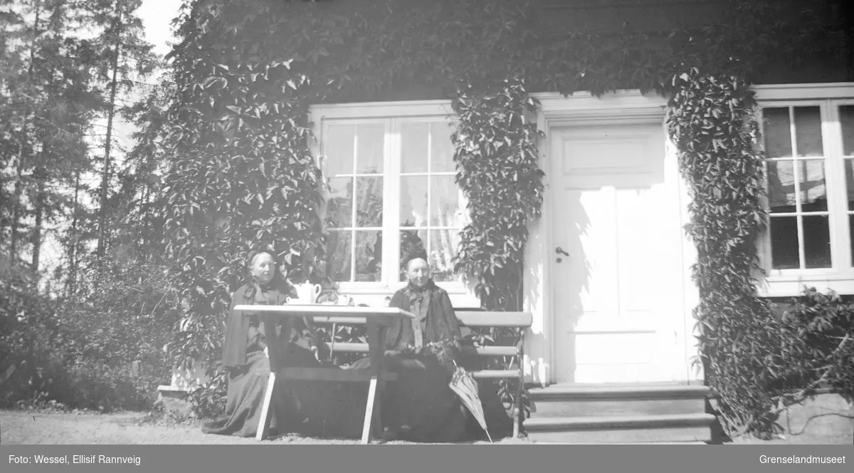 To kvinner fotografert sittende ved et bord utenfor et hus med mange slyngplanter, ukjent sted.