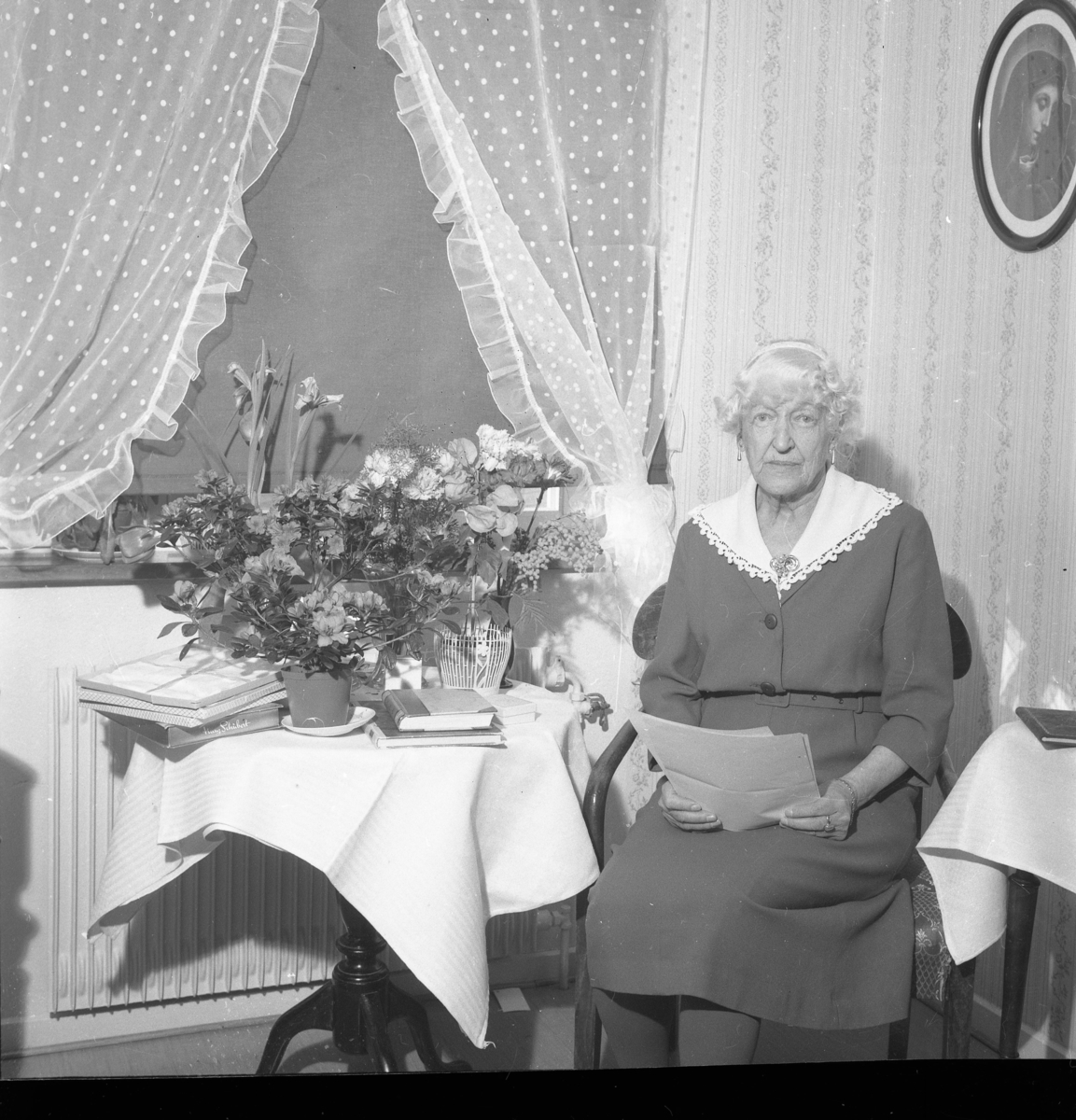Fröken Hård af Segerstad sitter vid ett bord med blommor och håller några papper i knät. Hon heter eventuellt Gertrud och fyller 80 år.