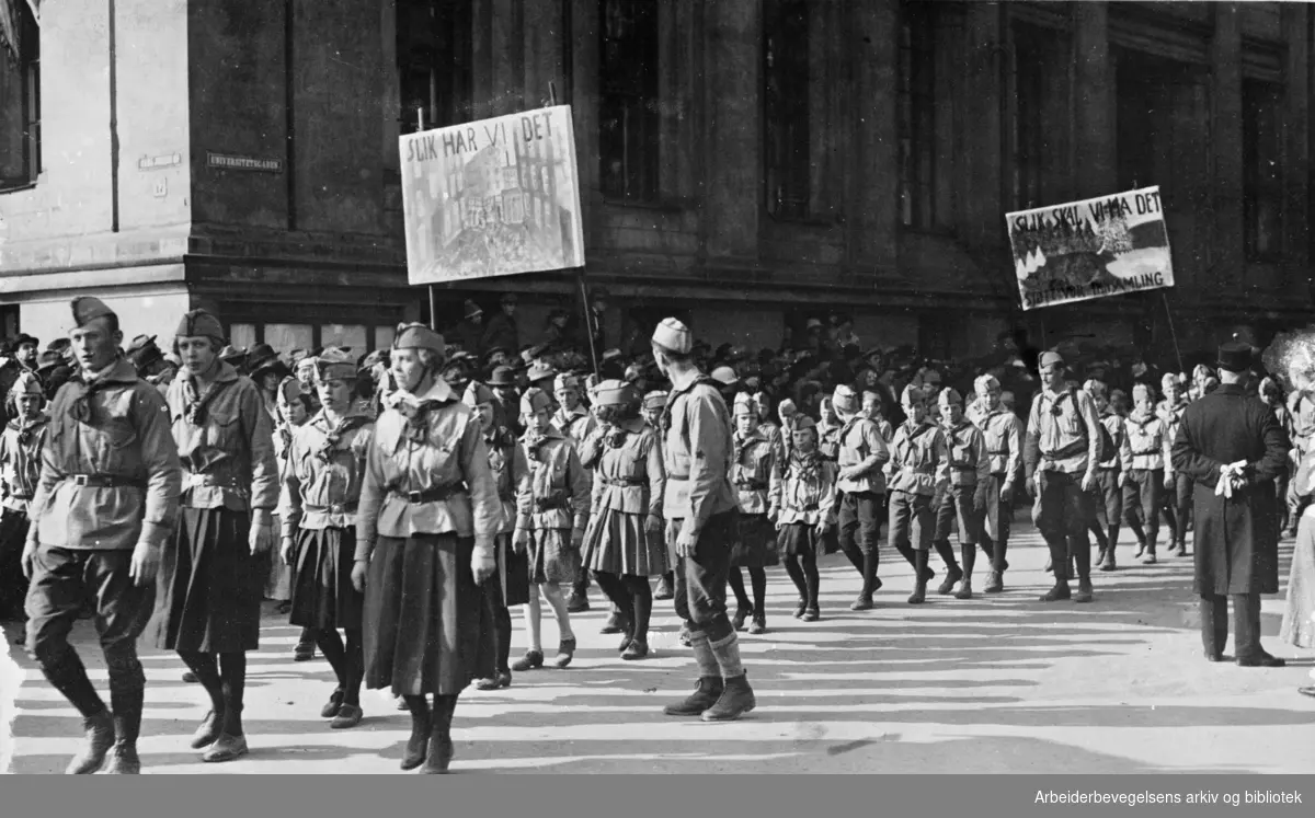 1. mai 1927. Røde speidere i demonstrasjonstoget som svinger inn på Karl Johans gate fra Universitetsgata. Paroler: Slik har vi det - Slik vil vi ha det/Støtt vor innsamling.