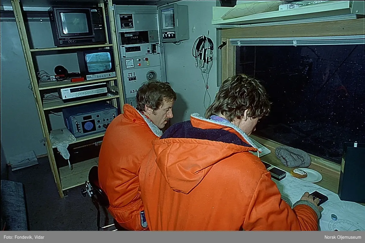 To menn ikledd orange kjeledresser studerer kart i et rom for dykkerkontroll (?)