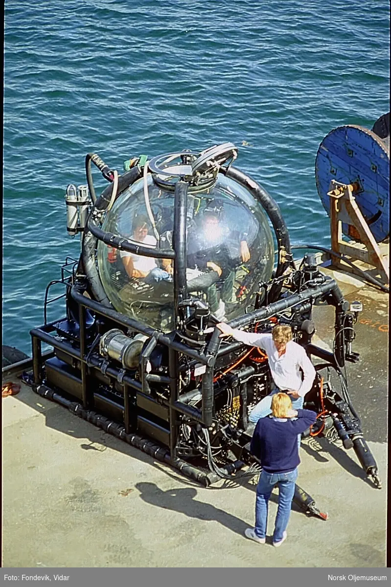 To personer står ved siden av miniubåten "Check Mate", mens to andre personer sitter inne i ubåten.