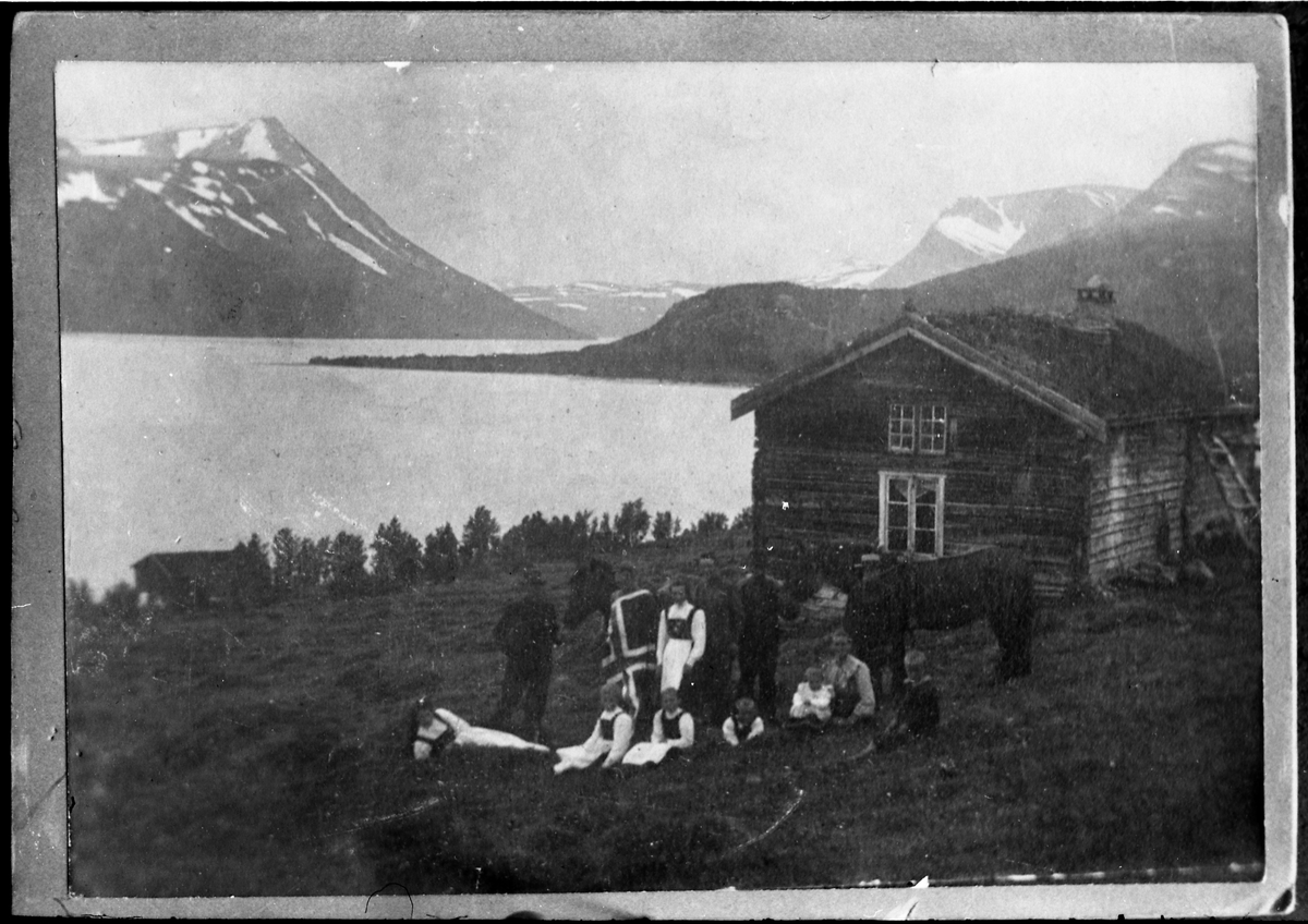 Avfotografert bilde som viser noen personer foran et tømret hus i fjelllandskap. I bakgrunnen en innsjø og fjell. Noen av personene holder et stort norsk flagg.