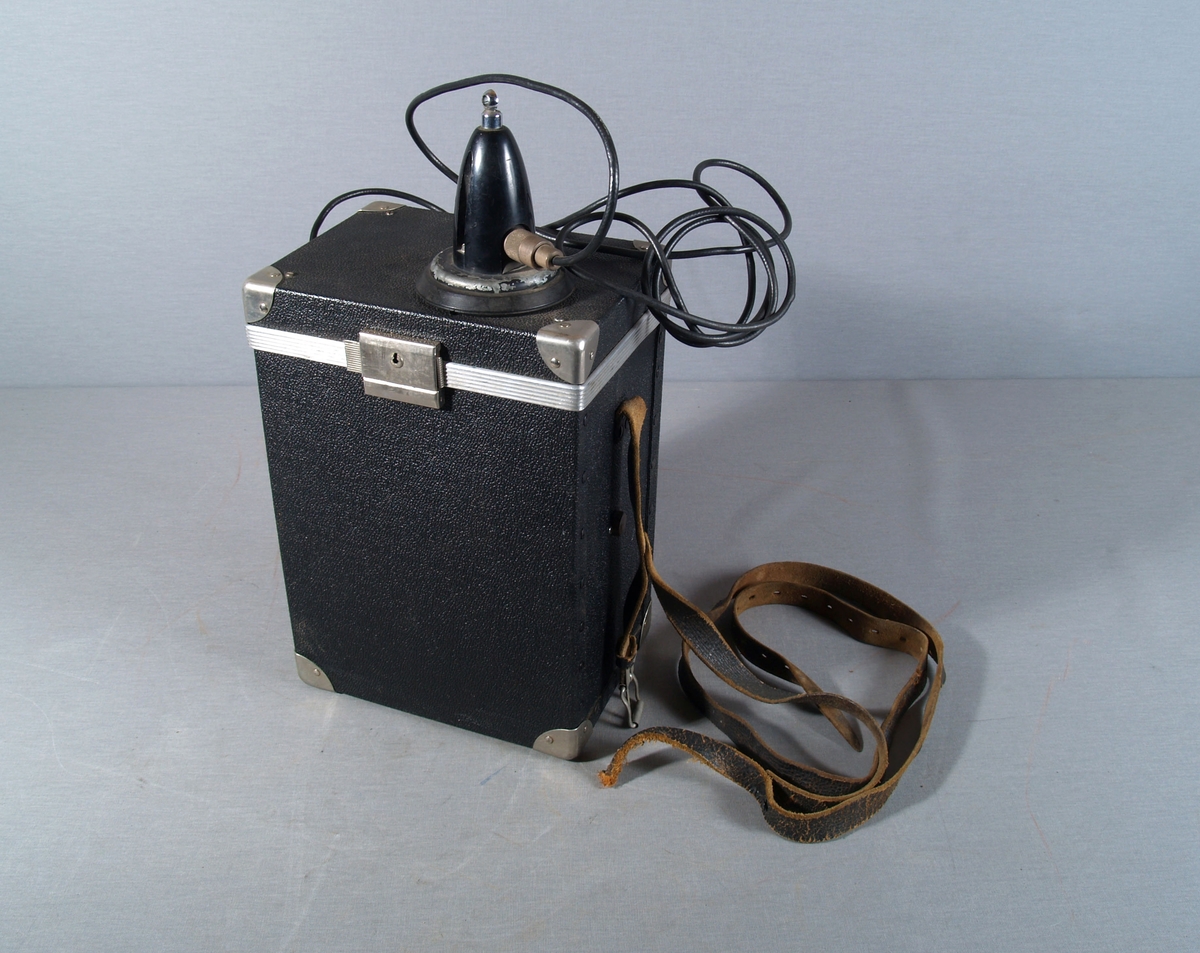 Bærbar radiotelefon med mikrofon i kasse med bærereim. Antennekabel (coax) ut gjennom kassens side og til antennebase/sokkel festet på kassens lokk.  Selve antennen mangler. Kassen har låsbart lokk.