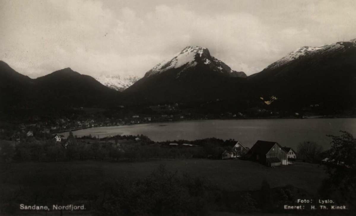 Postkort. Fotografisk motiv. Svart/hvitt. Motiv fra Nordfjord: Sandane. Landskap. Bebyggelse. Fjell. Fjord. Stemplet 14.09.1938.