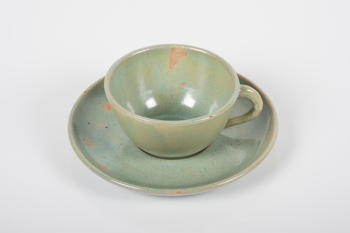 Kopp med skål i keramikk med grønn lasur. Buet hank på koppen. Skålen har spor etter tre knotter på bunnen, usikker funksjon. Bunnen på koppen og skålen har matt overflate.