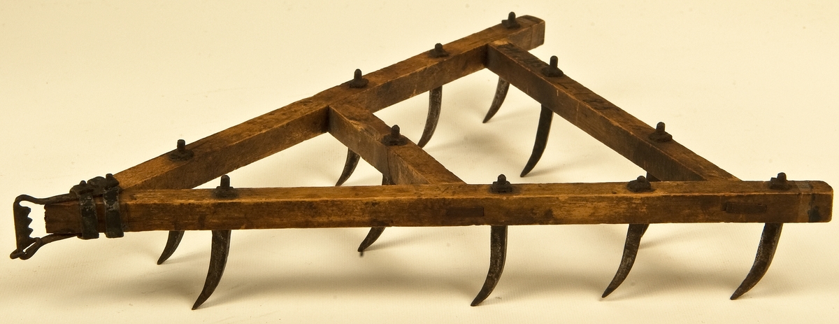 Modell till klösharv, åkerbruksredskap. Krokpinnharv. Trekantig av träbalkar med tvärslå på mitten. Elva stycken krökta järnpinnar. Längd 16 cm. Bredd 13 cm.