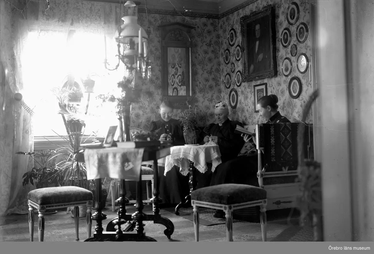 "Gamla hemmet", rumsinteriör, tre damer med handarbete.
Från höger Anna Thermaenius med mamma Jenny.
"Gamla hemmet" var det första som familjen bosatte sig i när man flyttade från Torshälla till Hallsberg 1868. Det började byggas av Johan Thermaenius.