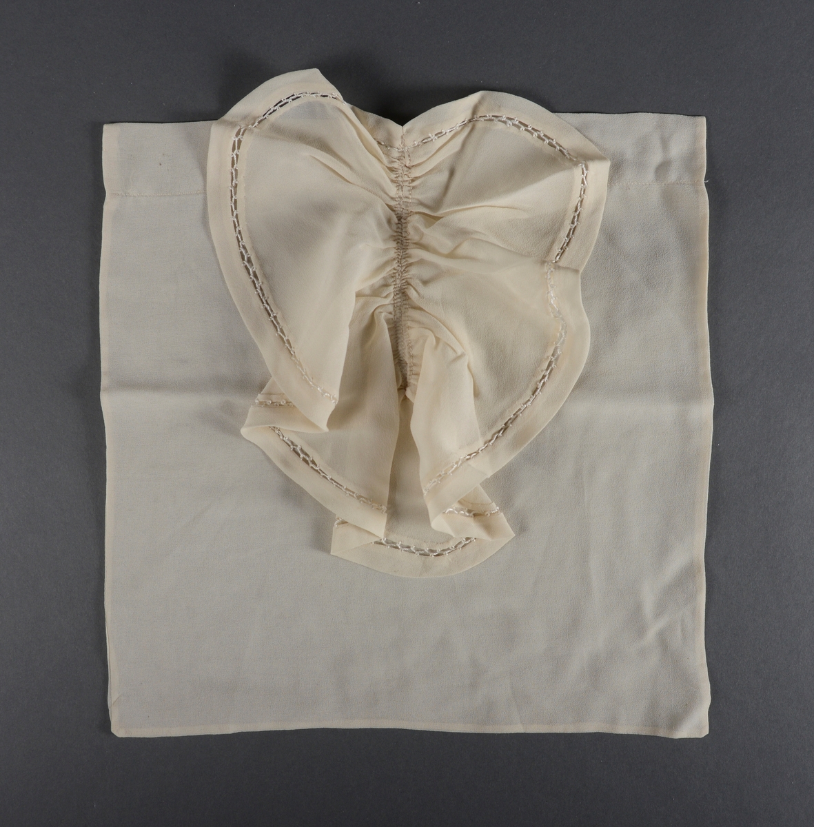 Et nesten kvadratisk skjortebryst laget av hvit chiffon. I front er det sydd på en volang som dekker nesten hele brystet. På volangen er det en heklet kant.