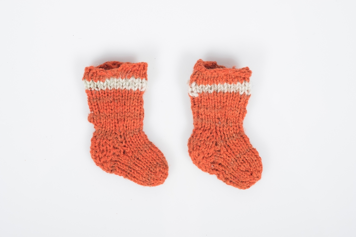 Et par bittesmå strikkede ullsokker. Sokkene er oransjerøde med en lys stripe øverst og nøen brunrøde striper lenger ned.