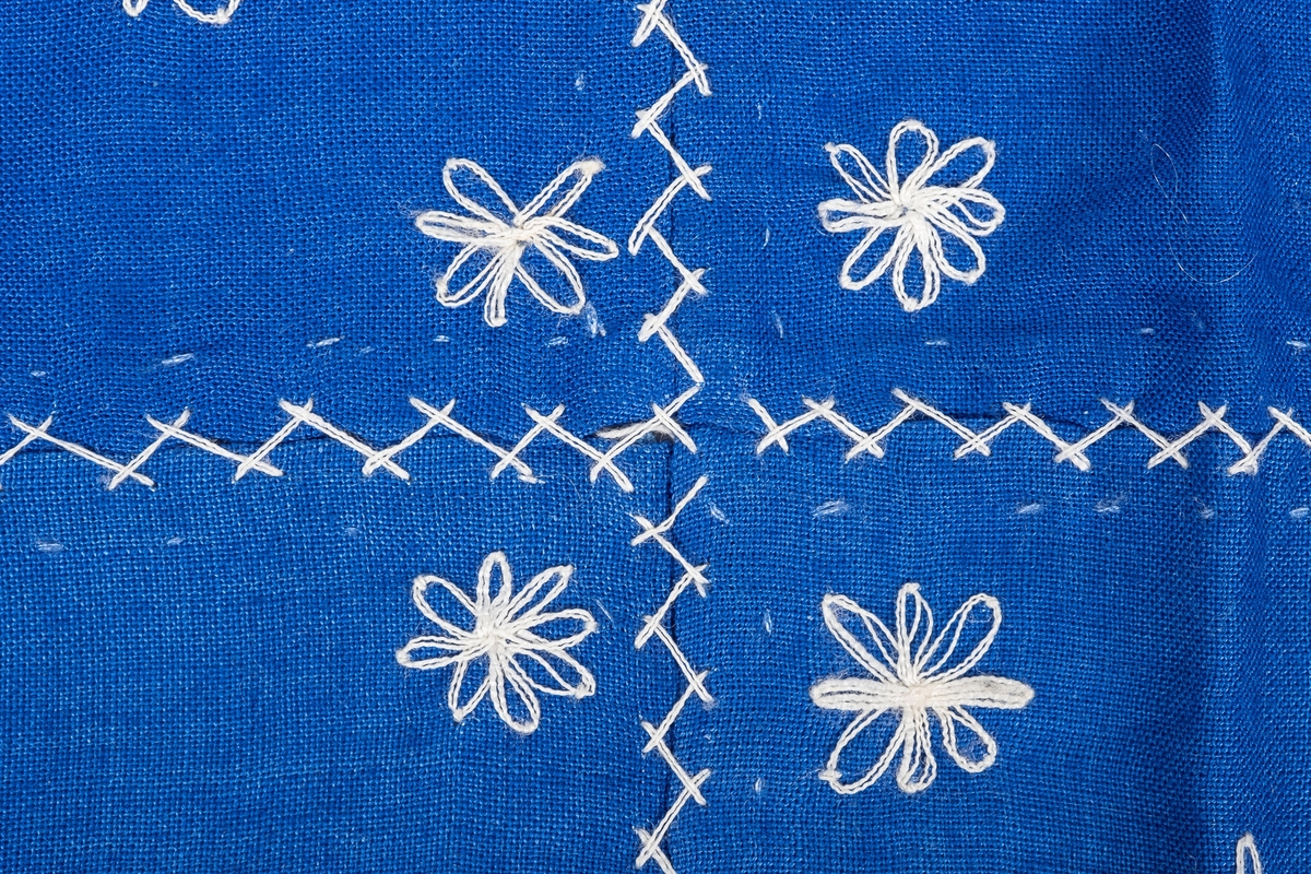 Nesten kvadraktisk kongeblått tøystykke med broderte små hvite blomster med 8 blomsterblader på. Brukt som vognteppe til dukkevogn. Satt sammen av 9 mer eller mindre kvadratiske tøybiter, sydd sammen med heksesting. Blomstene er gruppert i grupper på tre eller fem blomster per tøybit.