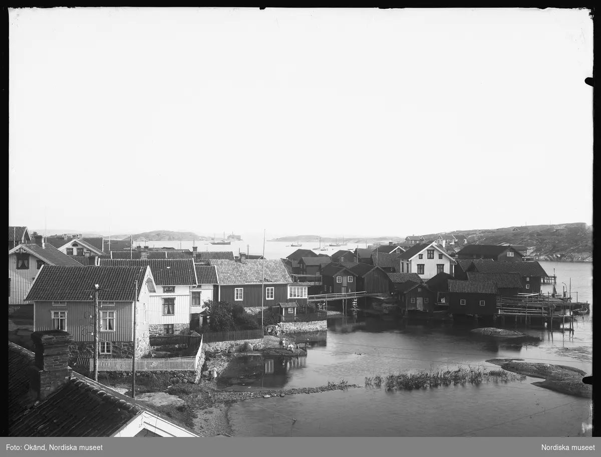 Mollösund, Orust Västra härad i Bohuslän 1921. Miljöbild över bebyggelse, strandlinje, bryggor och pirar med människor i rörelse. Bakom husen på udden skymtar flera båtar på den öppna vattenytan.  Västkusten