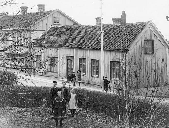 Två flickor och två pojkar står i trädgården till Brahegatan 60, snett mittemot Brahegatan 65 där andra barn står och tittar mot fotografen. En pojke står lutade mot en lyktstolpe.