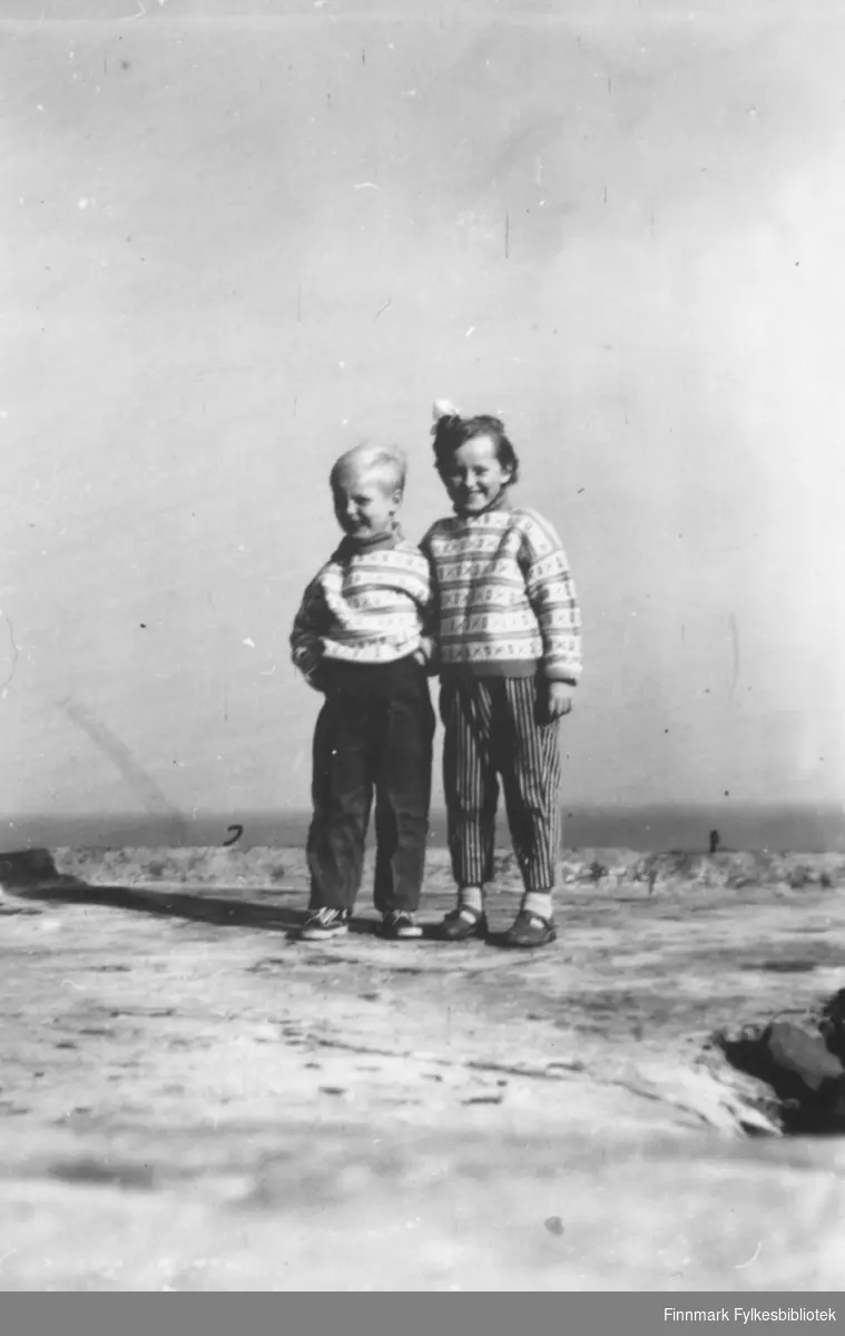 To barn fotografert i ullgensere, stående i sol og vind, muligens tatt på en strand eller elvebredde?  Familiealbum tilhørende familien Klemetsen. Utlånt av Trygve Klemetsen. Periode: 1930-1960.