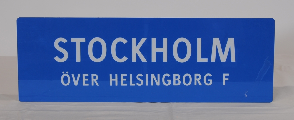 Destinationsskylt av ljusblå plast med texten "STOCKHOLM ÖVER HELSINGBORG F" i vitt. Spegelvänd text på baksidan.