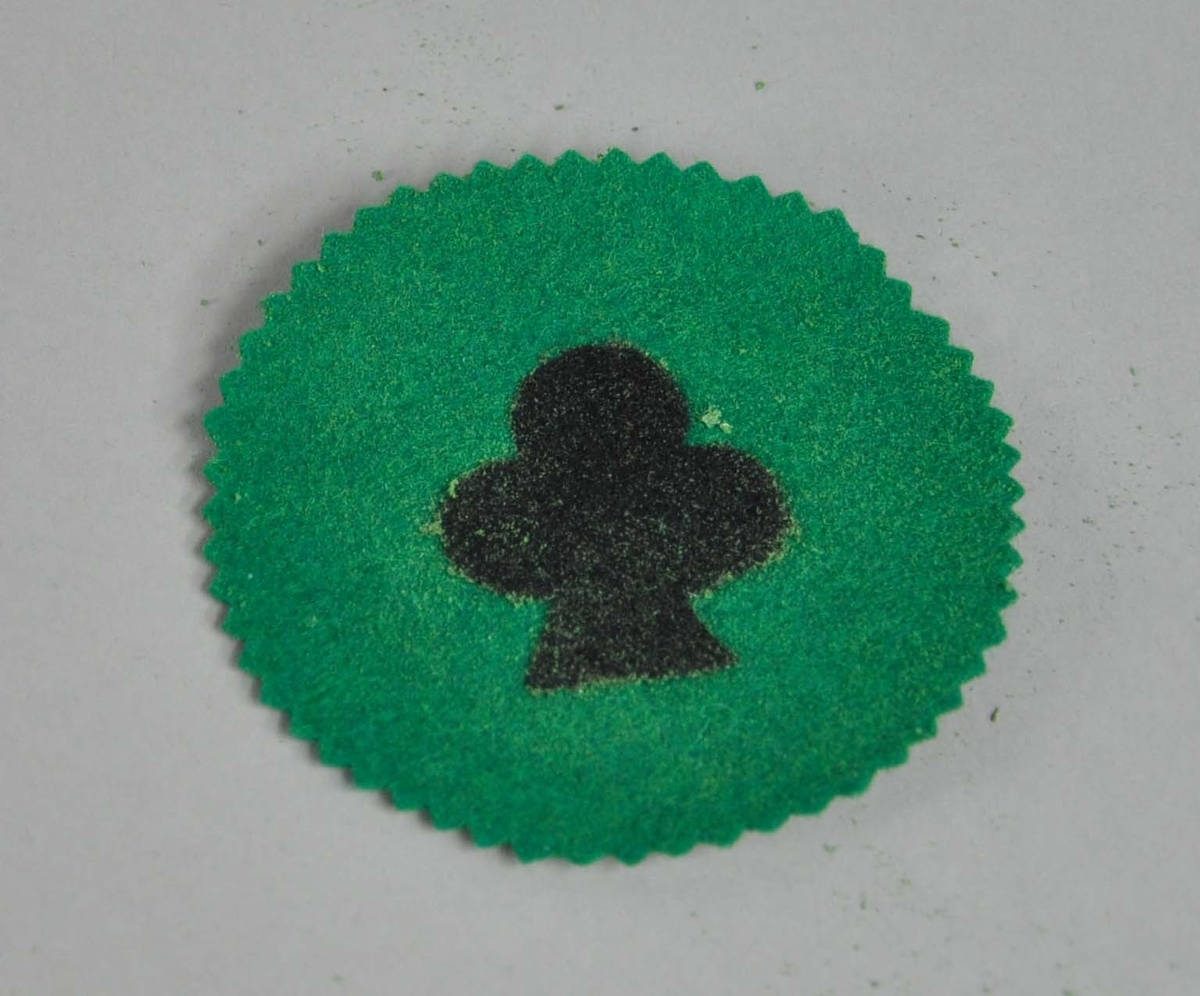 Rund brikke av grønt stoff, med bølgete kant. På midten er symbolet for kløverkortet.