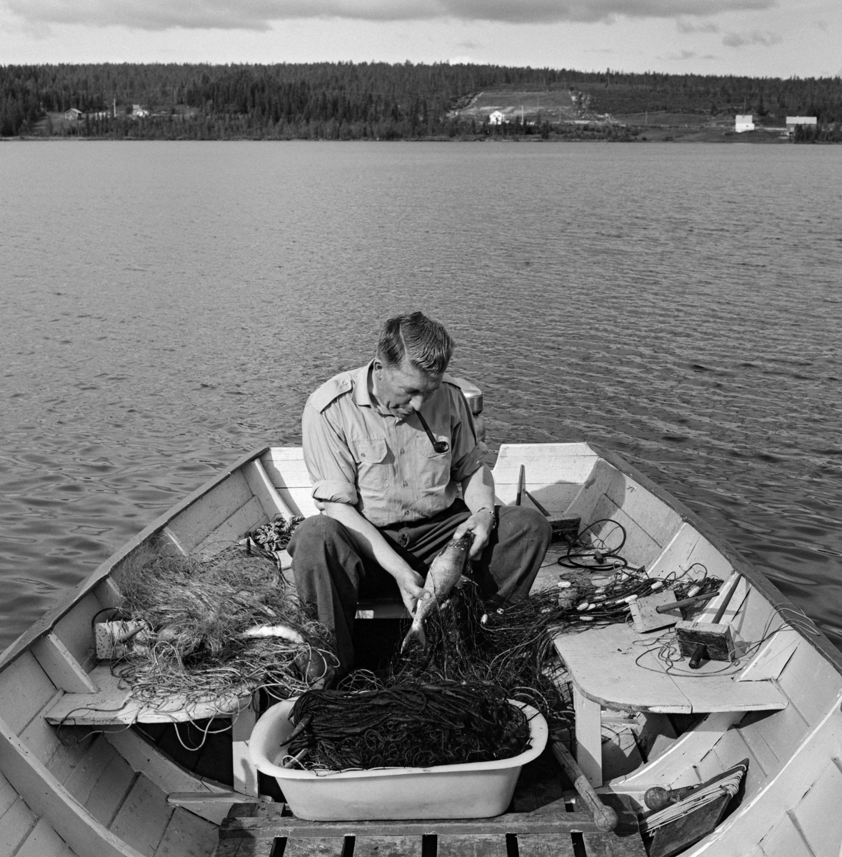 Per Lilleholm (1911-1999) fra Nordre Osen i Åmot kommune i Hedmark, fotografert i båten sin under garnfiske i Osensjøen.  Da dette fotografiet ble tatt satt Lilleholm på ei tofte akterut i båten sin og tok fisk ut av et garn som han la i ei plastbalje som sto på tiljene (golvdekningen) i småbåten av tre som han fisket fra.  Lilleholm var kledd i mørke bukser og ei lys skjorte med oppbrettete armer.  Han hadde tobakkspipa i munnen mens han trakk en forholdsvis stor sik ut av garnet.  I bakgrunnen ser vi det typiske skoglandskapet rundt Osensjøen, med et par gardsbruk i sonen ned mot vatnet. 