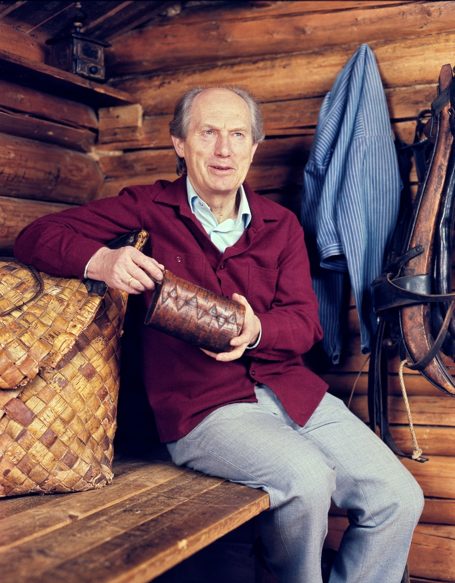 Museumsmannen Tore Fossum (1926-2017), fotografert i et koieinteriør i museets basisutstilling om skogbruk.  Bildet inngikk i en serie som ble tatt med tanke på Norsk Skogbruksmuseums årbok nr. 11 (1985-86), og som ble produsert som et festskrift i anledning Tore Fossums 60-årsdag.  Han var kledd i en burgunderrød busserull, lyseblå skjorte og grå bukser.  I hendene holdt han en neverbutt, mens han støttet den ene albuen på en kont.  På ei hylle oppunder raftet bak Fossum skimtes ei kaffekvern, og på veggen til venstre for ham hang det en busserull og en arbeidssele.  Museumslederen var plassert på kjørekarens plass i koia.  Tore Fossum ble født i Elverum i 1926.  Etter å ha tatt artium ved Hamar Katedralskole i 1945 gikk han blant annet Statens skogskole på Evenstad i Stor-Elvdal og Tekniska Skogs- och Sågverksskolan i Härnösand, før han i 1951 ble tatt opp som student på skogbrukslinja ved Norges Landbrukshøgskole.  Derfra ble han uteksaminert i 1954.  Etter et år ved Statens skogsforskningsinstitut i Stockholm ble han så ansatt som herredsskogmester i hjemkommunen Elverum.  I 1961 ble han tilbudt stillingen som konservator ved det da forholdsvis nyetablerte Norsk Skogbruksmuseum, som besto av 10-11 små husvær i friluftsmuseet på Prestøya og ei gjenstandssamling med snaut 500 objekter.  Konservatoren var den eneste ansatte.  Fossum tok fatt i utviklingsoppgaver på mange plan.  Da han gikk av som museumsdirektør ved Norsk Skogbruksmuseum i 1993 besto museet av et museumsbygg på cirka 12 000 kvadratmeter fordelt på tre plan, et friluftsmuseum med om lag 40 antikvariske bygninger i Elverum og 9 på filialavdelingen Sørlistøa fløtermuseum.  Museet hadde store samlinger av gjenstander, fotografier og arkivalia.  Fossum engasjerte seg også i kulturhistorisk dokumentasjon og forskning, og var pådriveren bak 12 årbokutgivelser og en rekke kulturhistoriske dokumentasjonsfilmer.  For denne innsatsen ble han tildelt kongens fortjenstmedalje i gull i 1993.  Som pensjonist bisto Tore Fossum museet med ledelse av filialavdelingen Sørlistøa fram til og med 2001, og han engasjerte seg i Elverum historielag, der han var redaktør for årboka «Alfarheim» fram til 2008.