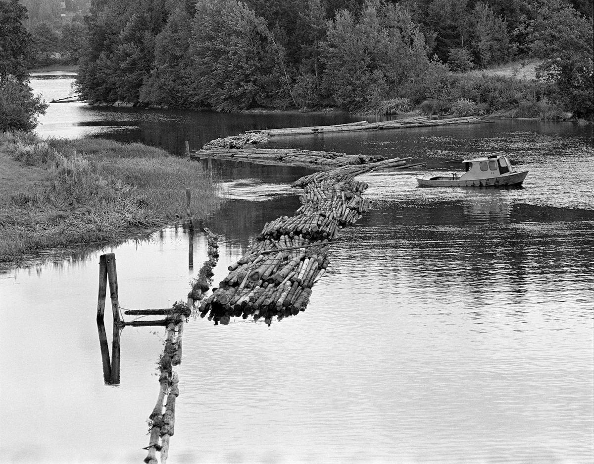 Buntet fløtingstømmer ved innløpet til Strømsfoss lense i Aremark i Østfold. Fotografiet er tatt i 1982, den siste sesongen det ble fløtet tømmer i Haldenvassdraget.  I sluttfasen ble det utelukkende fløtet ubarket massevirke i tre meters lengder.  Tømmeret ble levert i lastebilbunter, som ble samlet i store slep over de mange innsjøene i vassdraget.  Ved slusing måtte imidlertid slepene deles opp i kortere lenker som fikk plass i slusekamrene.  Av det nevnte sortimentet gikk det fire bunter i hver slusevending.  Ved oppdeling av slepene ovenfor slusestedene og sammenbinding nedenfor ble slepene buksert med små varpebåter av stål, som den vi ser på dette fotografiet.  Haldenvassdragets Fellesfløtningsforening hadde 18 slike.  Fartøyet på fotografiet het «F8» - F-en refererte til Fellesfløtningsforeningen.  Bildet viser også at tømmeret lå fortøyd ved ei dobbelt ledelense, som var lagt slik at endene på lensestokkene overlappet hverandre, som takstein.  Bildet viser også hvordan lensa var støttet av en duc d’albe, en pælebukk, i et sumpete elvelandskap.

En liten historikk om tømmerfløting og kanaliseringsarbeid i Haldenvassdraget finnes under fanen «Opplysninger».
