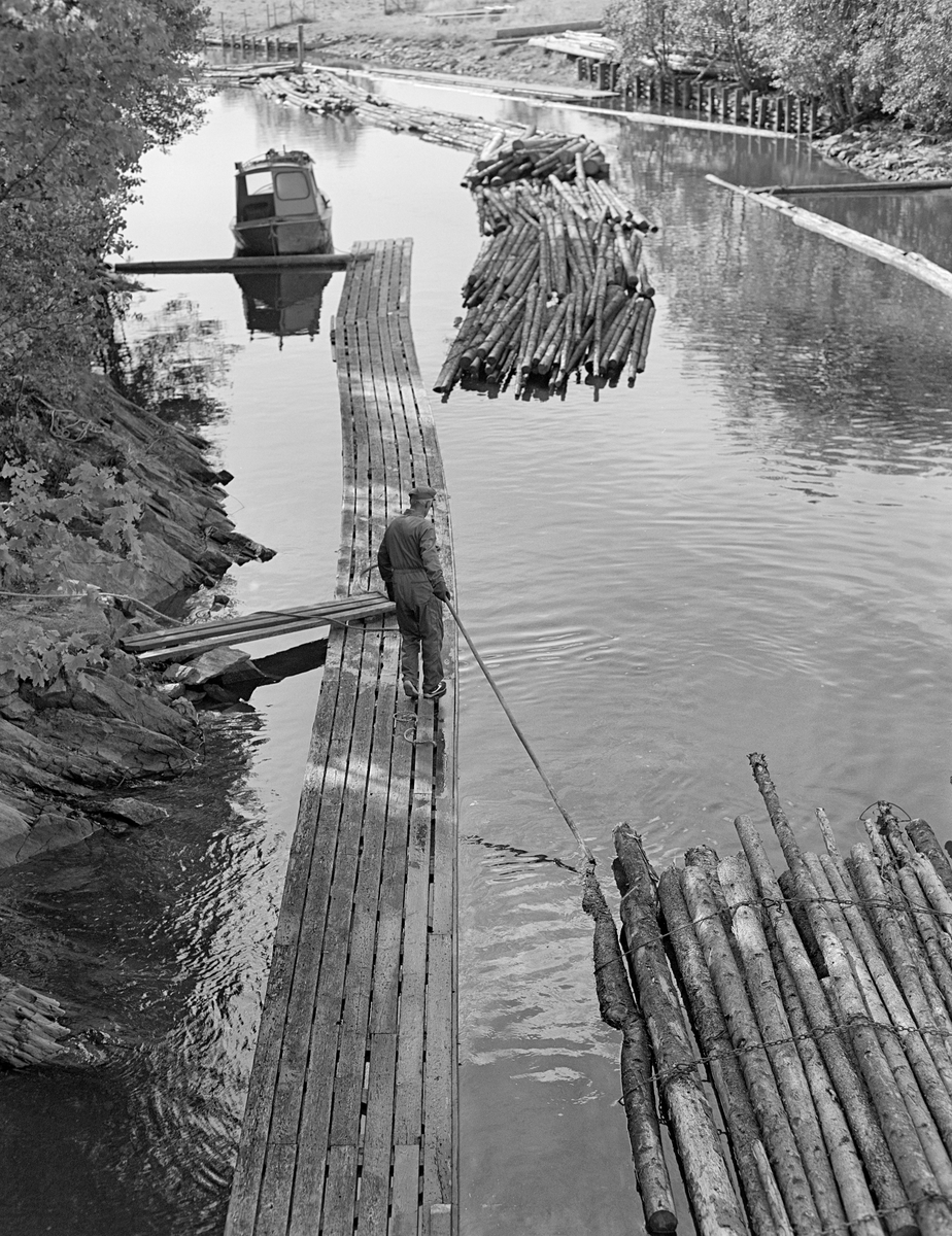 Fra kanalen nedenfor Strømsfoss sluser i Aremark kommune i Østfold.  Fotografiet er tatt i 1982, som var den siste sesongen det ble fløtet tømmer i Haldenvassdraget.  Vi ser fra plattformen ved nedre sluseport mot en gangbane, der det gikk en kar med en langskaftet fløterhake.  Haken brukte han til å trekke den fremste bunten i ei slusevending som nettopp hadde passert sluseanlegget ned mot overenden av ei lenke av bunter som hadde kommet ned tidligere.  Buntene skulle kjedes sammen, ende mot ende, i ei lang lenke.  Seinere skulle flere slike lenker bindes sammen til større innsjøslep.  Ved enden av gangbanen en varpebåt av stål som ble brukt under arbeidet med slepene.

En liten historikk om tømmerfløting og kanaliseringsarbeid i Haldenvassdraget finnes under fanen «Opplysninger».