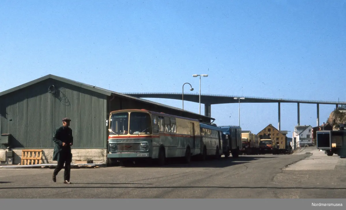 Langs havneskuret står en rekke godsvogner som kan identifiseres. Fremst står UX13061, en kombinertbuss som gikk i rute mellom Stramsnes og Kristiansund. Det var Peder og Nils Anton Strømsvåg drev disse rutene. UX13061 ble registrert våren 1972. I 1975 ble Strømsvåg sine overtatt av Kristiansund-Oppdal Auto (KOA) og bussen ble etter kort tid solgt til private eiere. Det var derfor få år denne bussen gikk i rute på Kristiansund.
Bak denne ses to vogner det ikke er lett å identifisere. Men det synes å ha blått tak, slik som vognene til KOA. Bak disse to står et vogntog i rødt og gult, mest sannsynlig et som gikk for Surnadal Billag. (Info: Sveinung Berild). -  havneskur på Storkaia i sentrum på Kirkelandet i Kristiansund. I bakgrunnen ser vi Sørsundbroen. fra samling av Fotografier og karttegninger, fotografert eller samlet sammen av Arkitekt MNAL Kristian Sylthe (f. 18.06.1926) ved Kristiansund kommune i sitt virke som kommunearkitekt (senere næringssjef) samt som sekretær i Oljeutvalget fra begynnelsen tidlig 1970. Samlingen kan i hovedsak dateres mellom 1945 til 1980. Samlingen er gitt i gave av Kristiansund kommune. Fra Nordmøre museums fotosamlinger.